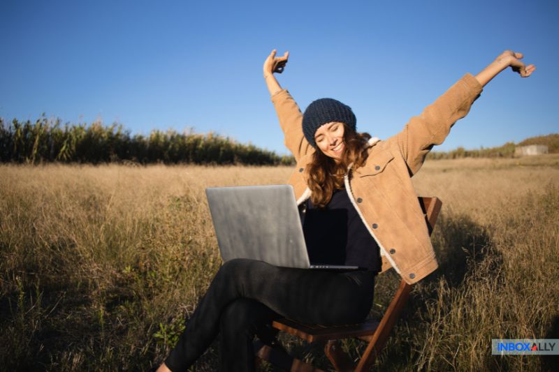 A person sits outdoors on a chair with a laptop smiling at the boost in cold email open rates, all while enjoying a grassy field under a clear blue sky.