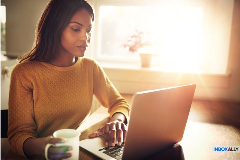 A woman works on a laptop while holding a mug, pondering if cold emailing works