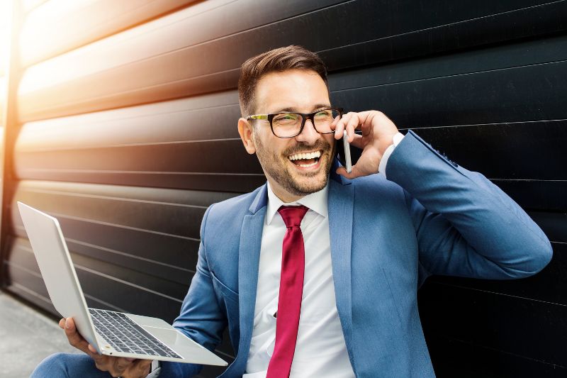 Um empresário sorridente, de óculos, segurando um laptop enquanto discute estratégias de e-mail frio ao telefone