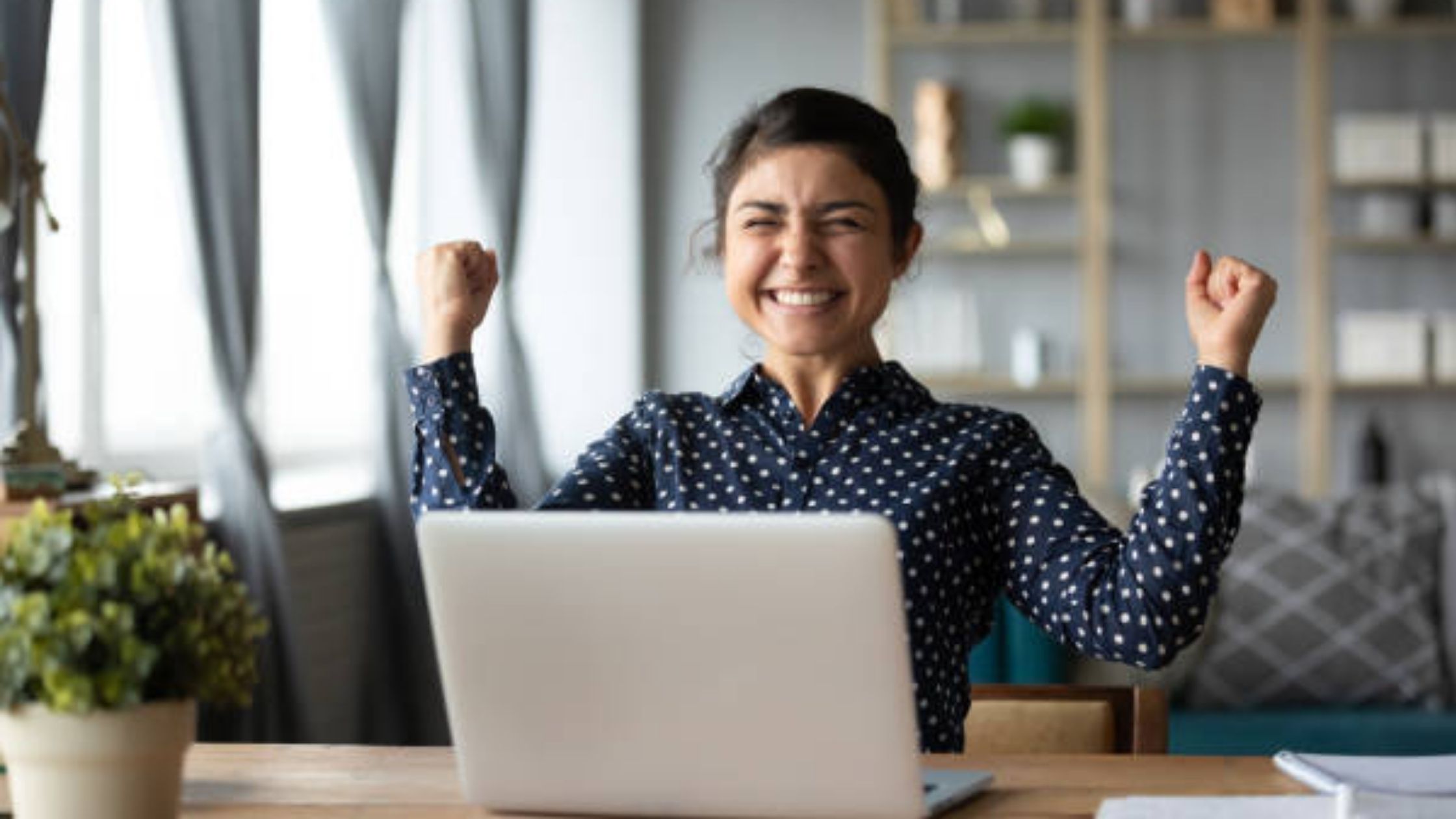Uma mulher comemora com os punhos erguidos em frente ao seu laptop em uma mesa de escritório em casa depois de aprender a escrever e-mails frios com sucesso.