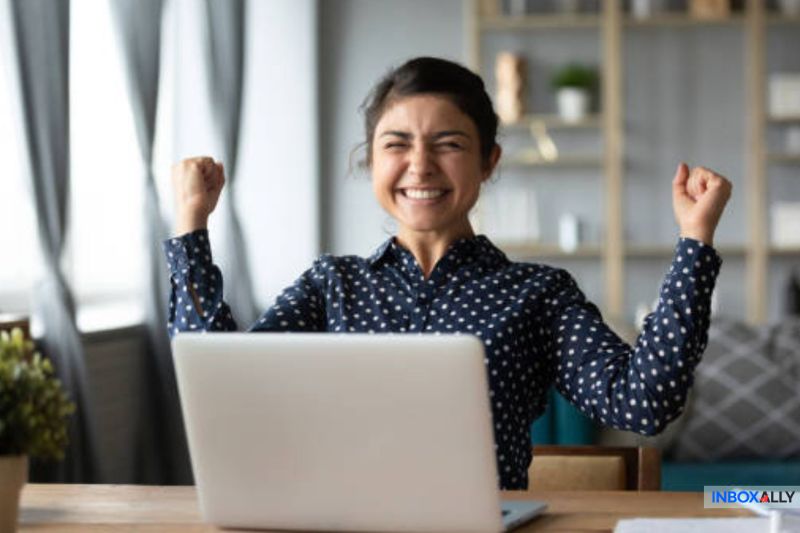 A person sits at a desk, smiling with arms raised in excitement, in front of an open laptop, celebrating the success of cold emails that get opened.