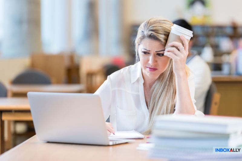 A woman looks stressed as she holds a coffee cup, contemplating strategies for crafting cold emails that get opened while sitting at her laptop in a library or office setting.