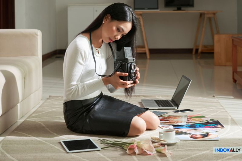 A woman sits on the floor examining a camera, contemplating her next shot. Around her are printed photos, a tablet displaying strategies for cold emails that get opened, a laptop, a cup of coffee, and flowers.