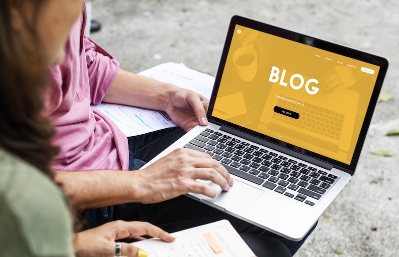 Two people sit at a table outdoors, one holding notes while the other types on a laptop displaying a blog page, likely discussing the best guest post email templates 