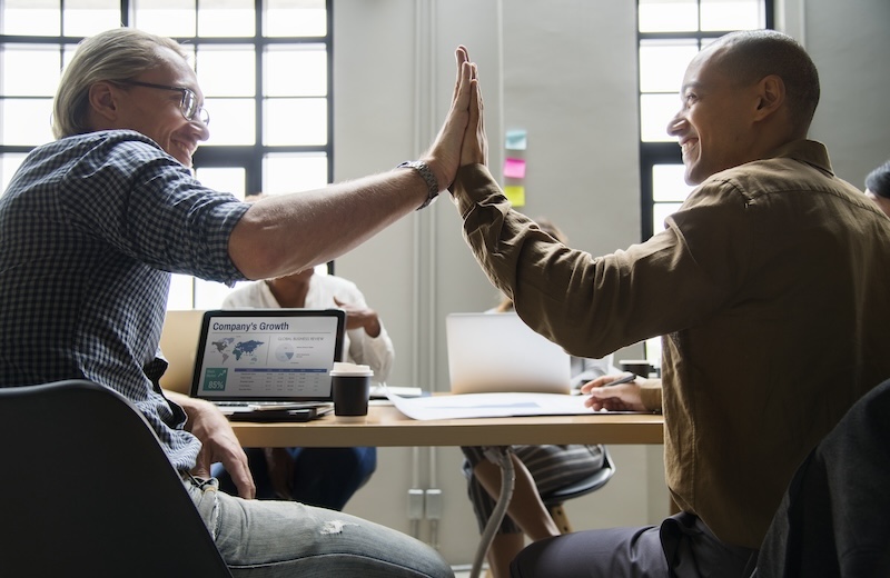 Duas pessoas sorrindo e dando um high-five em uma reunião de escritório. Um laptop exibindo um relatório de 