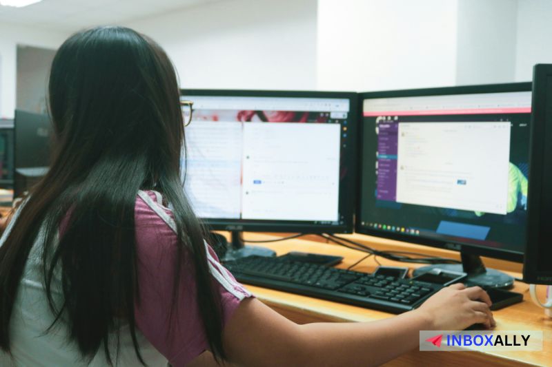 A person works at a desk with two computer monitors displaying various software applications; the rectangular InboxAlly logo, hinting at mass email management.