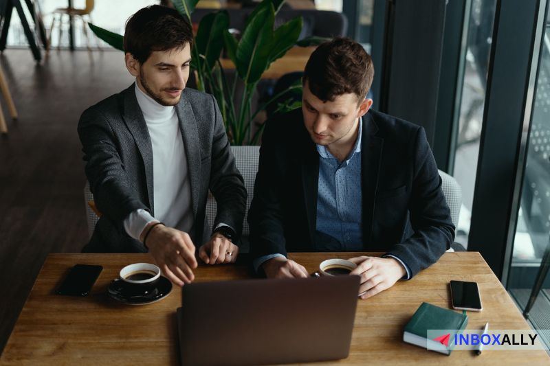 Two men in business attire sit at a wooden table, engaging in a discussion about the best LinkedIn outreach strategies while looking at a laptop. 