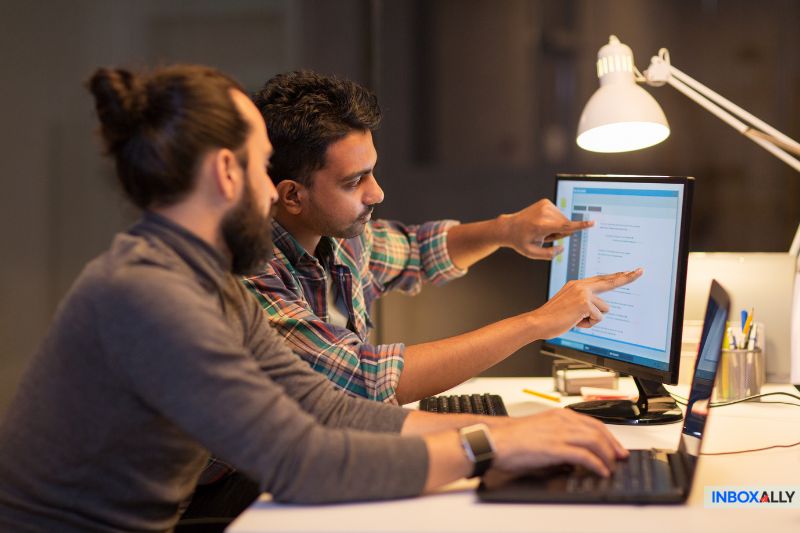 Duas pessoas colaborando em uma mesa, apontando para uma tela de computador que exibe texto sob uma lâmpada de mesa, exploram estratégias de growth hack.