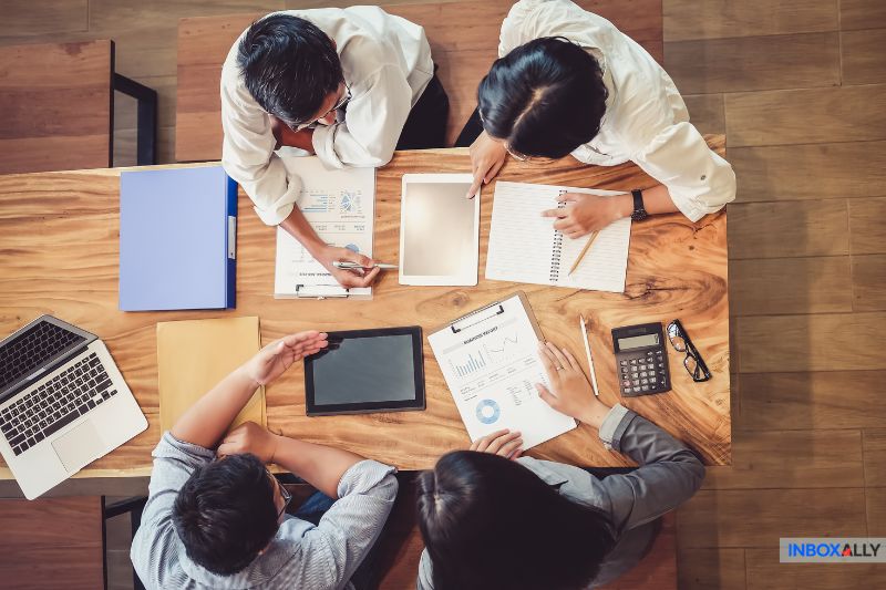 Quatro pessoas se sentam ao redor de uma mesa de madeira com laptops, tablets, papéis, uma calculadora e um notebook, envolvidos em uma animada discussão sobre estratégias de growth hack e técnicas de apostas de reputação.