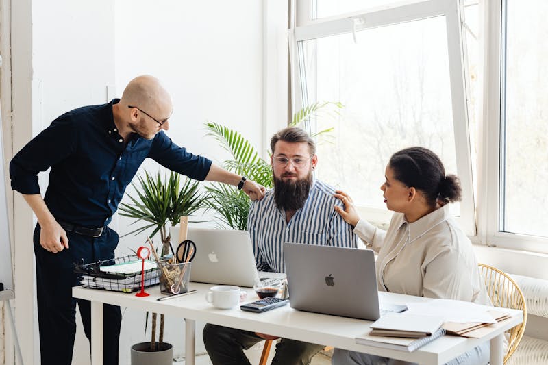 Three co=workers are gathered around a desk with laptops in a bright office. One person stands pointing while the other two sit, focusing on how to build trust. As they engage in discussion on crowded inbox