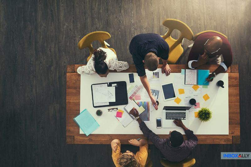 Aerial view of six people collaborating around a wooden table with laptops, notepads, and documents, strategizing their next email marketing campaign.