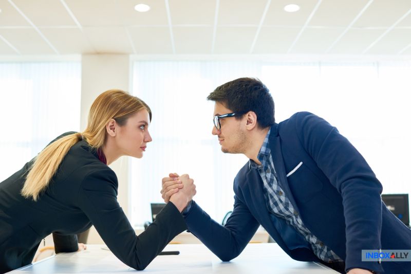 In the office setting, a man and a woman in business attire engage in arm wrestling across the table, embodying the spirit of competitive analysis.