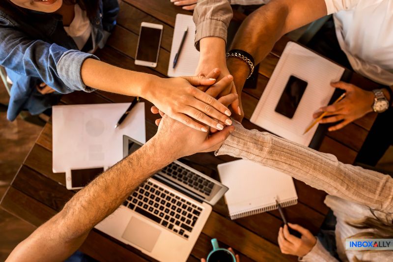 The group engages in a collaborative effort, placing their hands together over a table filled with notebooks, phones, and pens, as they dive into an intense session of competitive analysis.