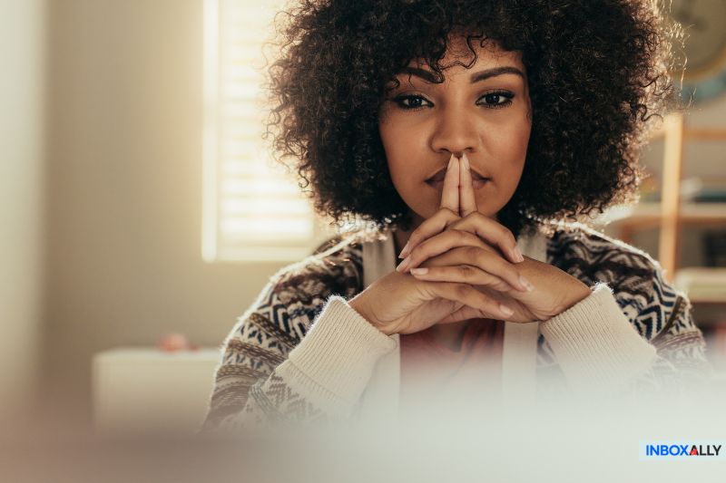 Uma pessoa de cabelos cacheados está sentada com os dedos pressionados, parecendo pensativa. Ela está usando um suéter estampado, iluminado suavemente ao fundo, como se estivesse refletindo sobre as nuances da elaboração de e-mails de marketing eficazes.